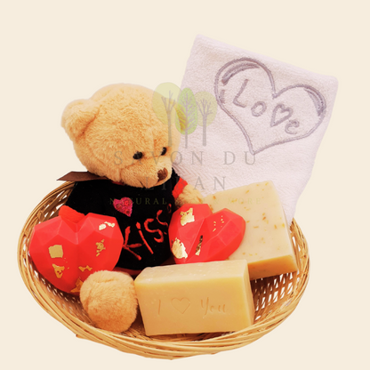 Love Gift Set "Tes Caresses" made of a round straw basket including a Teddy Bear, 2 Heart Soaps, 1 Embroidered Towel, 1 Oat Scrub Soap and 1 Musk Soap stamped with "I ❤️ You".